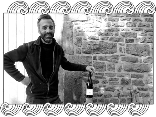 La Maison Romane, Burgundy winemaker, holding a bottle against a stone wall, framed with Dynamic Cellars wave design