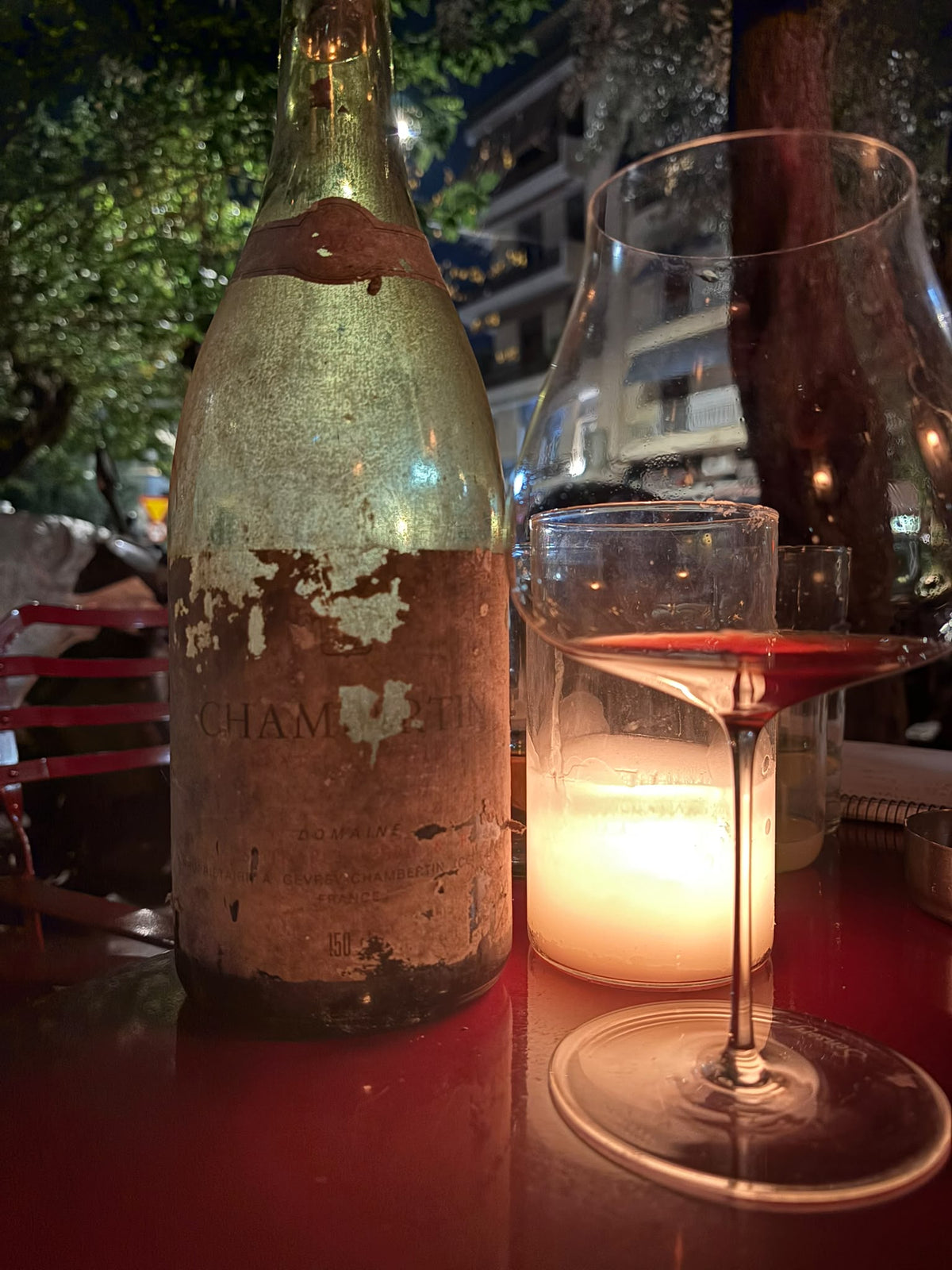 An old magnum of Chambertin with a weathered label, beside a glass of red wine and a candle on a terrace at night